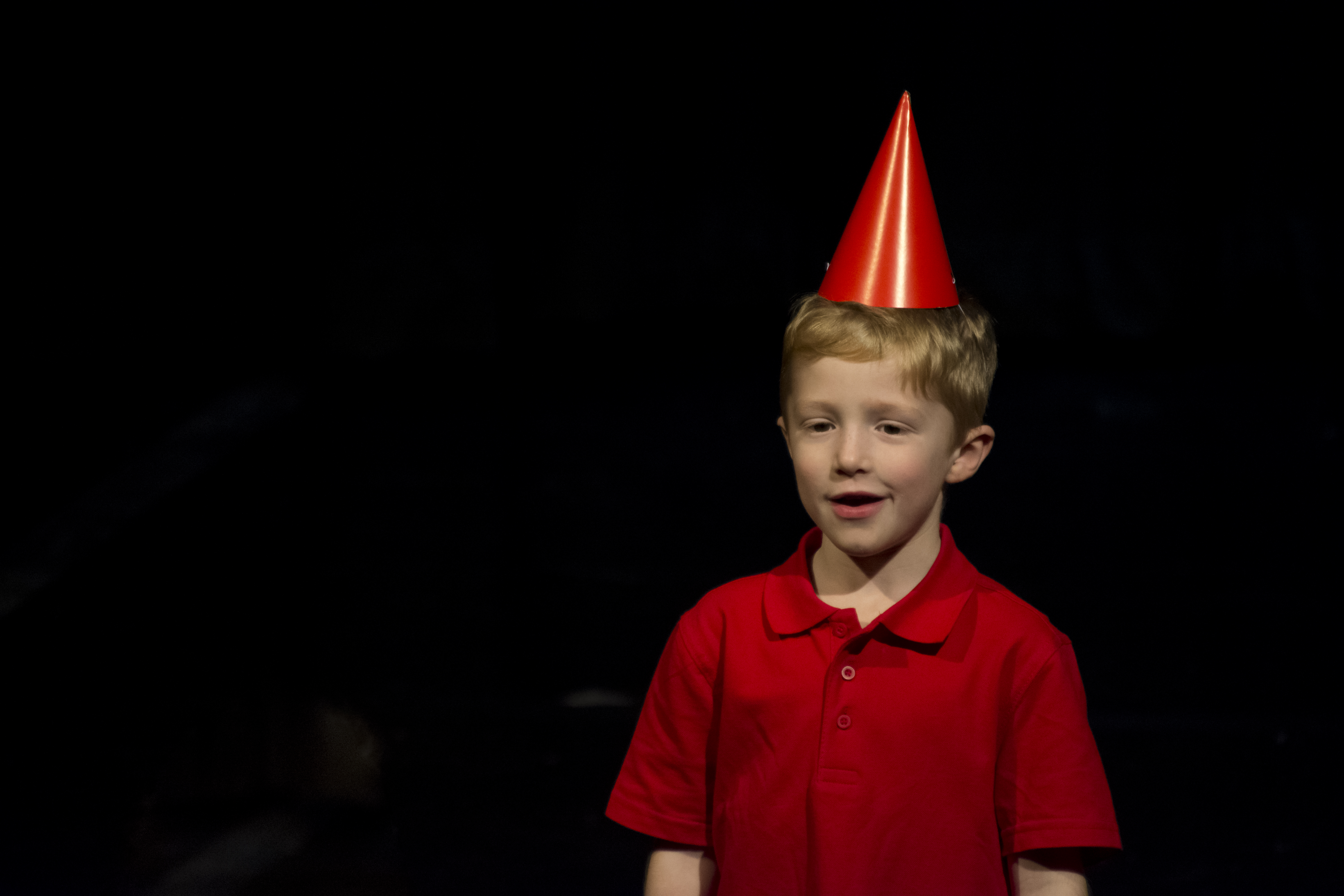 Young boy in a red shirt and party hat performing on stage.